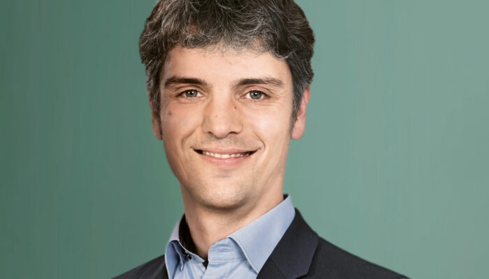 Ein Mann im Anzug lächelt vor einem schlichten grünen Hintergrund.