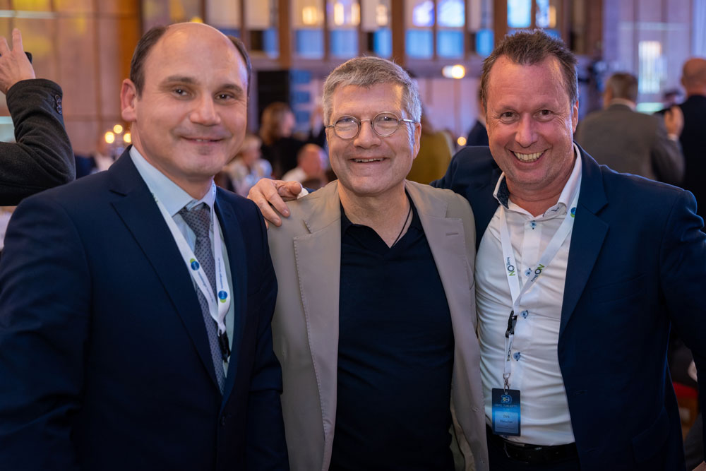 Drei Männer in Geschäftskleidung lächeln gemeinsam bei einer Veranstaltung oder Konferenz in einem Gebäude.