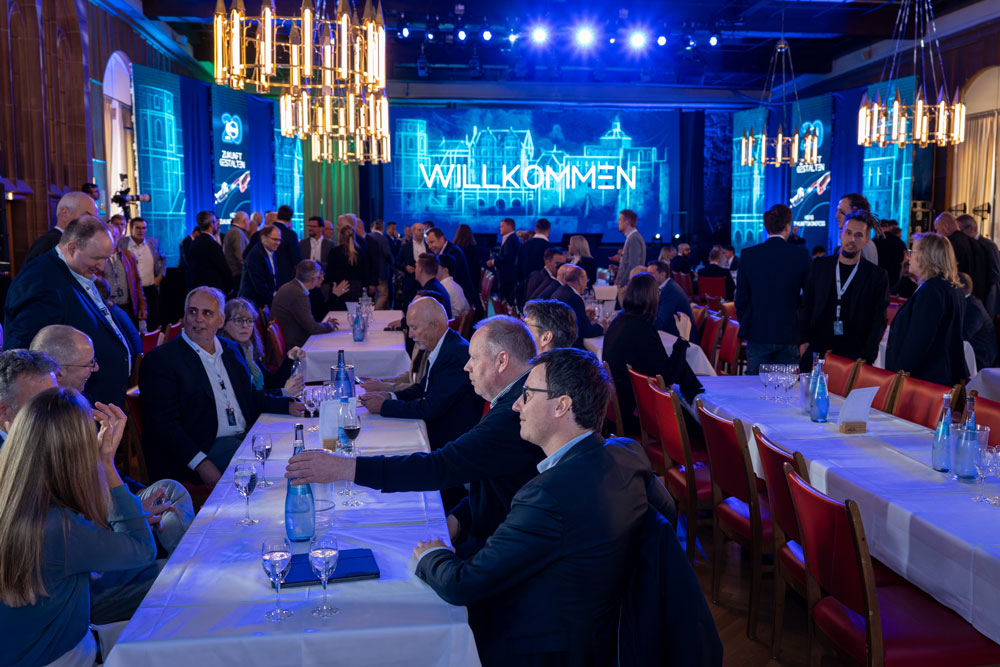 Die Menschen versammelten sich in einer großen Halle, in der vorne auf einer Leinwand "Willkommen" angezeigt wurde.
