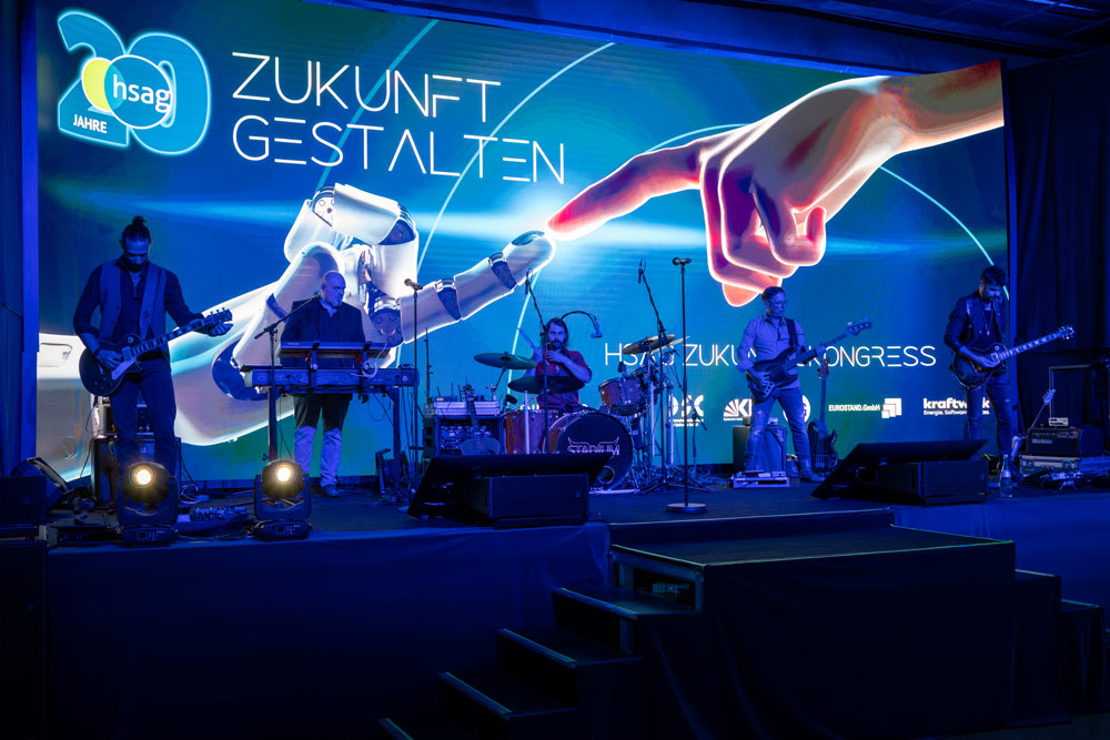 Eine Band tritt auf der Bühne vor einem futuristischen Bildschirm mit einer Roboterhand und deutschem Text auf.