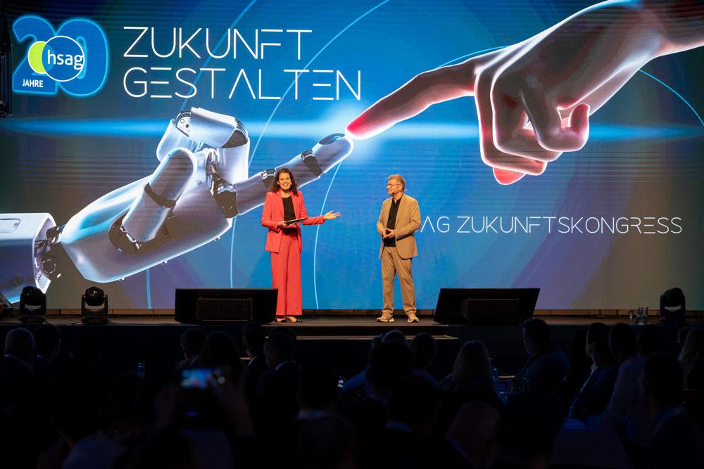 Zwei Sprecher auf der Bühne mit einem großen Bildschirm, der eine Roboterhand zeigt, die auf eine menschliche Hand zeigt.
