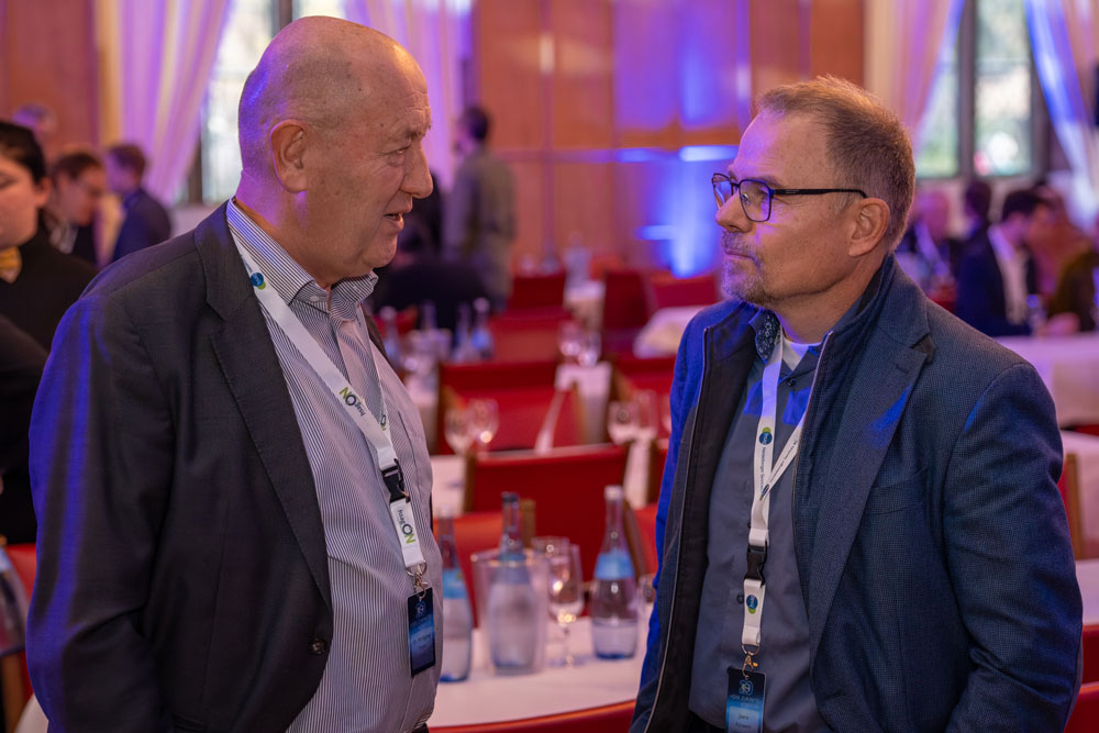 Zwei Männer im Geschäftsanzug unterhalten sich auf einer Konferenz mit Menschen und Tischen im Hintergrund.