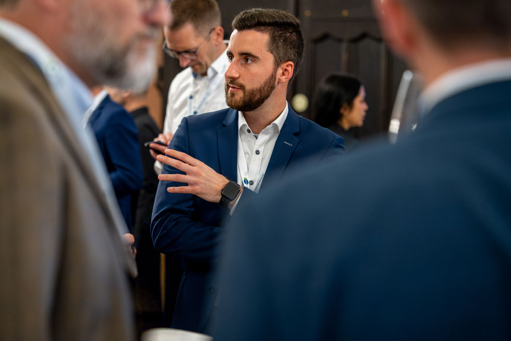 Ein Mann im Anzug spricht bei einem Networking-Event, andere Personen sind im Vorder- und Hintergrund unscharf.