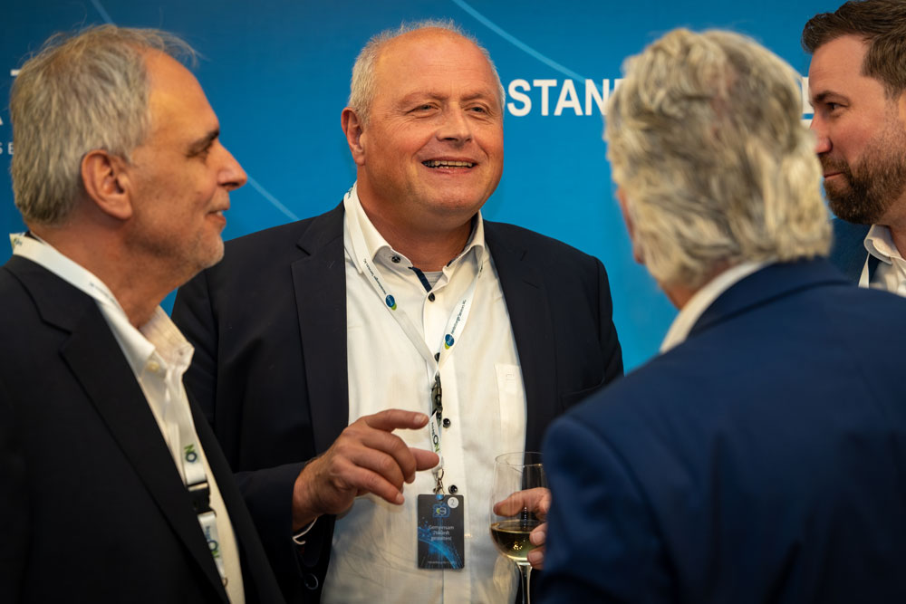 Vier Männer in Anzügen unterhalten sich und lächeln bei einer beruflichen Veranstaltung, einer hält ein Glas Wein in der Hand.