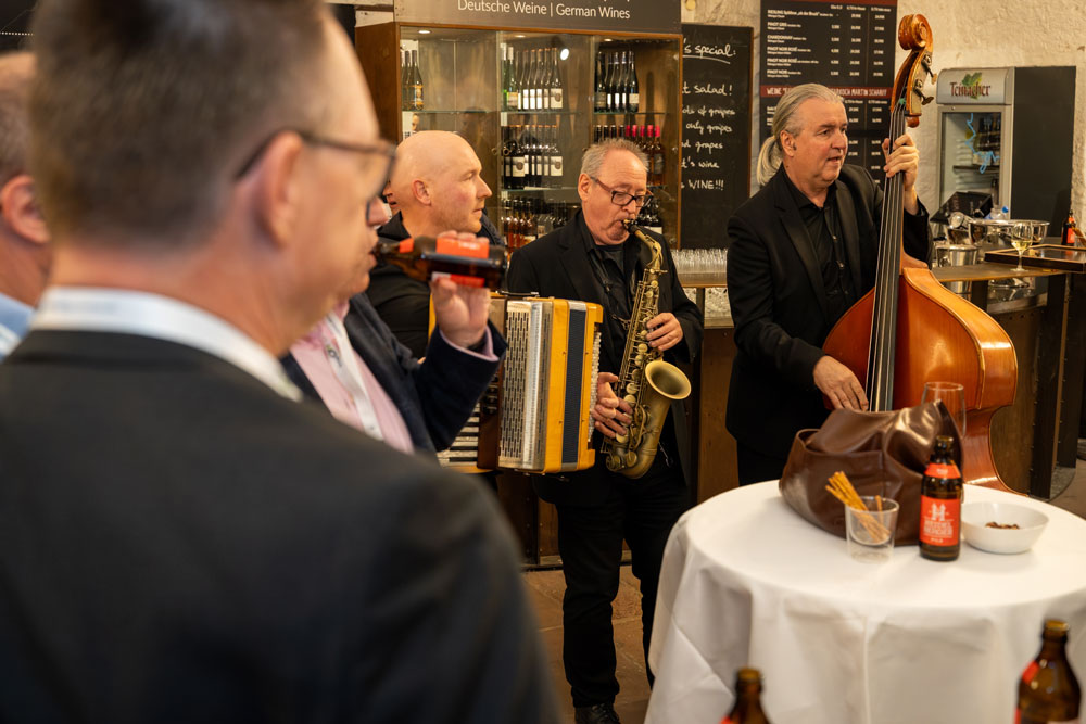 Eine kleine Jazzband spielt bei einer Indoor-Veranstaltung neben einem Tisch mit Getränken und Snacks.