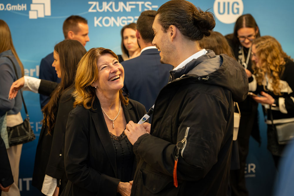 Zwei Personen lachen und unterhalten sich bei einer beruflichen Veranstaltung mit anderen im Hintergrund.
